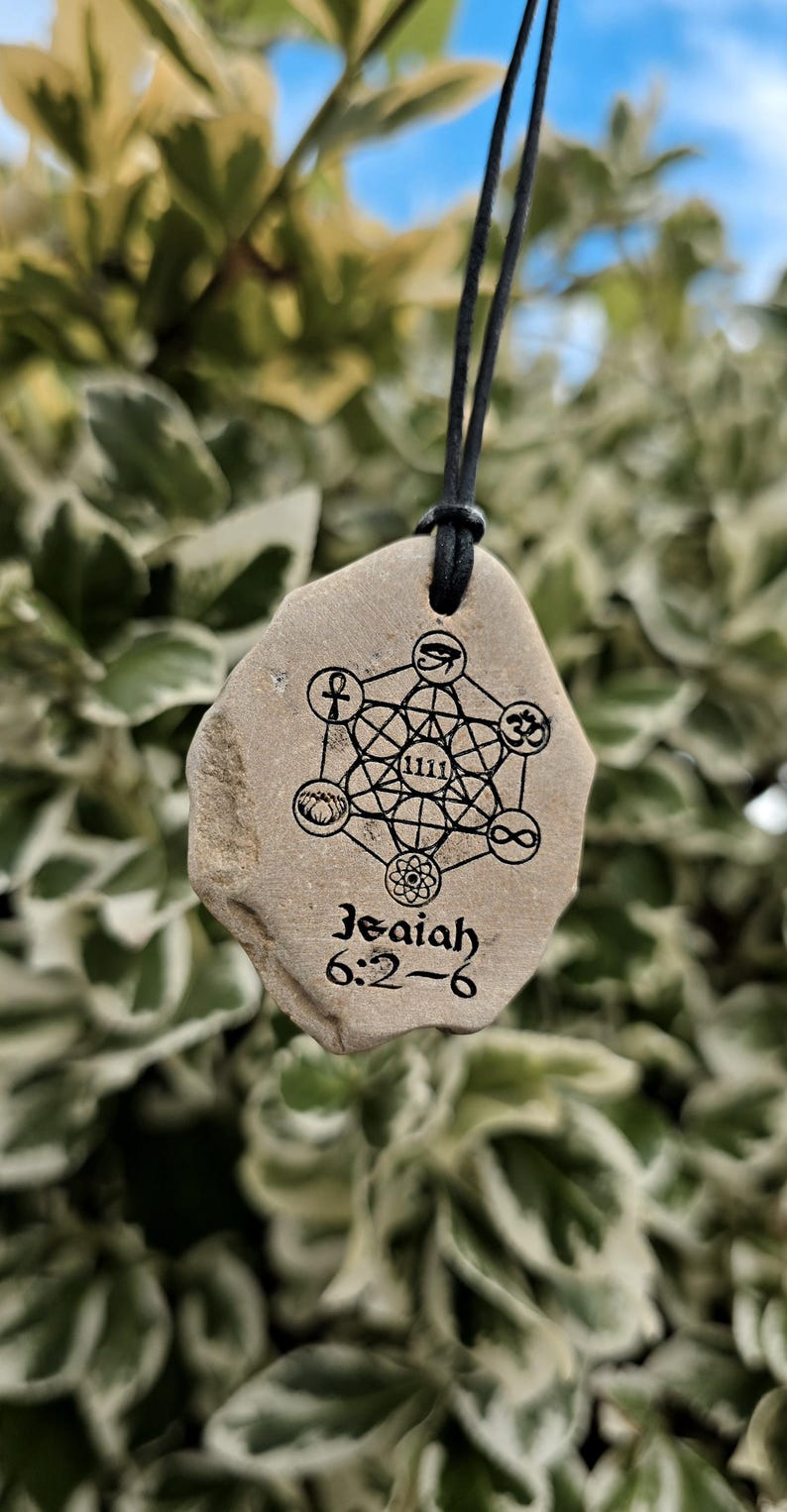 May include: A tan, irregularly shaped stone pendant hanging from a black cord. The stone features a black printed design of Metatron's Cube with various symbols, the number 1111, and the text Isaiah 6:2-6 printed in a black serif font.