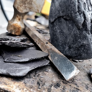 May include: A weathered metal hammer with a yellow handle, a long flat metal chisel, and several thin, dark gray slate stone fragments arranged near a large, rough-textured, dark gray stone block on a wooden surface.