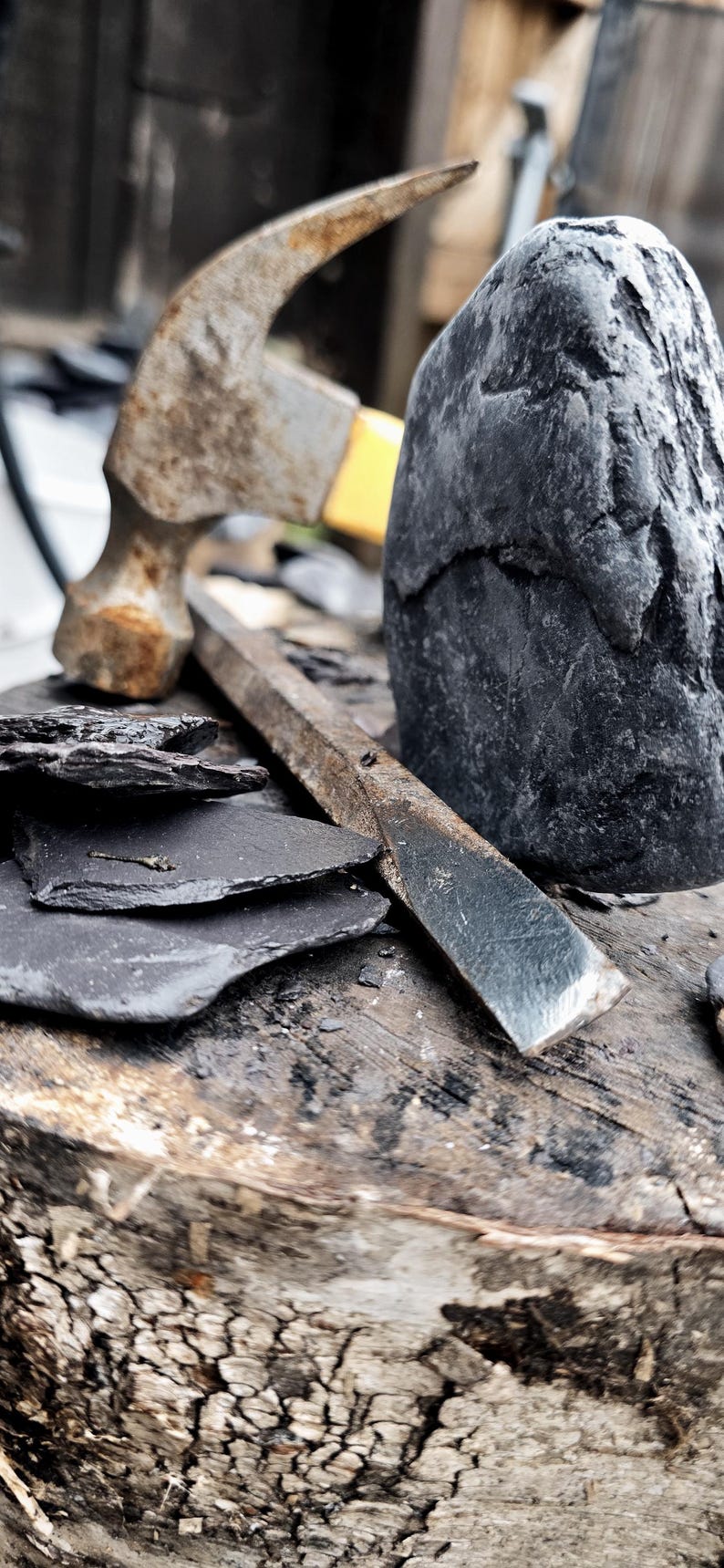 May include: A weathered metal hammer with a yellow handle, a long flat metal chisel, and several thin, dark gray slate stone fragments arranged near a large, rough-textured, dark gray stone block on a wooden surface.