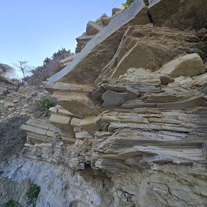 May include: A steep, natural rock formation featuring layered sedimentary strata in shades of tan, beige, and gray. The cliff face displays jagged, protruding stone plates and weathered textures, with small patches of green vegetation visible.