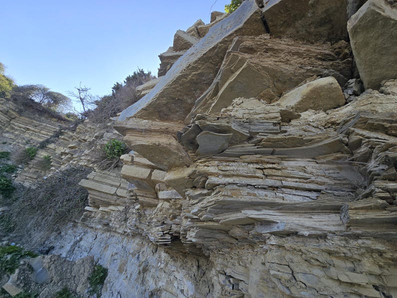 May include: A steep, natural rock formation featuring layered sedimentary strata in shades of tan, beige, and gray. The cliff face displays jagged, protruding stone plates and weathered textures, with small patches of green vegetation visible.