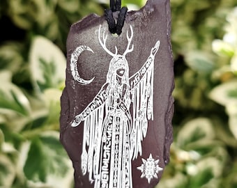 May include: A dark grey, irregularly shaped stone pendant hanging from a black cord. The stone features a white engraved illustration of a figure with antlers, wearing a long robe with fringe, standing between a crescent moon and a star.