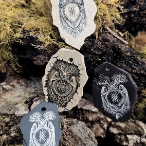 May include: Four irregular, flat stone pendants with pre-drilled holes at the top. Each pendant features a detailed line art illustration of two hooded figures standing beneath a tree, surrounding a central geometric star symbol in black or white.