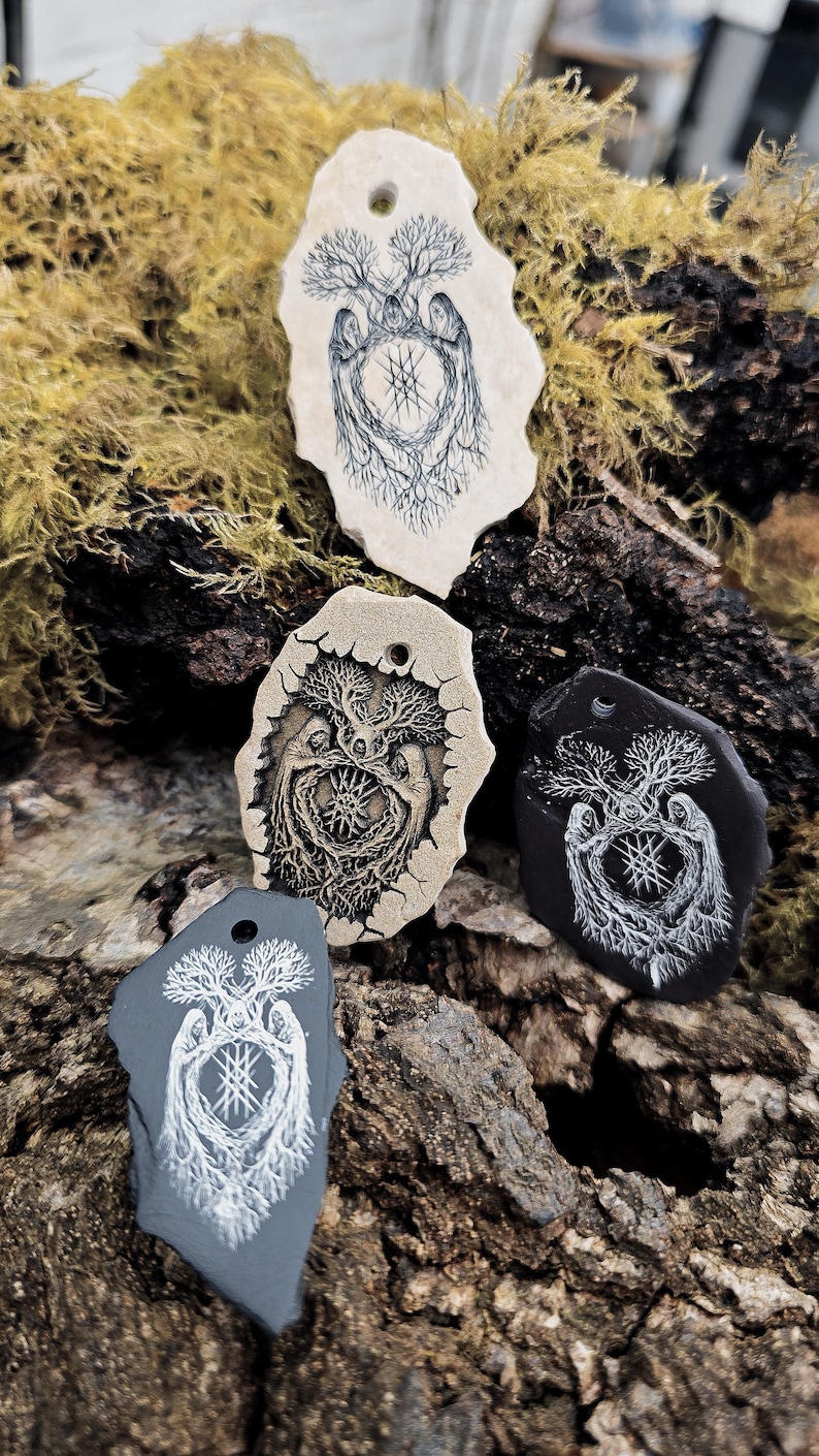 May include: Four irregular, flat stone pendants with pre-drilled holes at the top. Each pendant features a detailed line art illustration of two hooded figures standing beneath a tree, surrounding a central geometric star symbol in black or white.