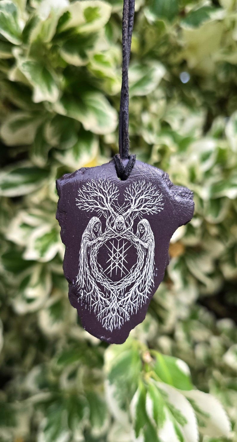 May include: A dark purple, irregularly shaped pendant hanging from a black cord. The surface features a white line-art illustration of three hooded figures standing in a circle, surrounded by intricate tree branches and roots with a central geometric symbol.