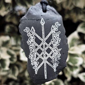May include: A black, irregularly shaped stone pendant suspended from a black cord. The surface features a white, hand-engraved design of a central vertical spear crossed by two diagonal spears, all intertwined with detailed leafy vine patterns.