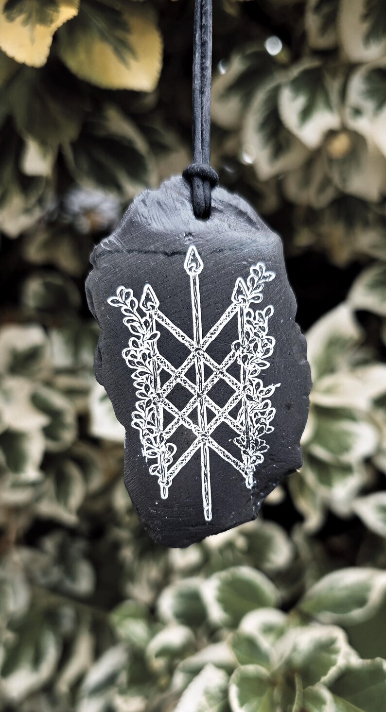 May include: A black, irregularly shaped stone pendant suspended from a black cord. The surface features a white, hand-engraved design of a central vertical spear crossed by two diagonal spears, all intertwined with detailed leafy vine patterns.