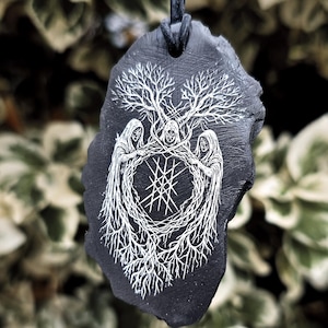 May include: A black slate pendant suspended on a dark cord, featuring a white engraved illustration of three hooded figures standing in a circle, their robes merging into intricate tree roots and branches surrounding a central geometric star symbol.