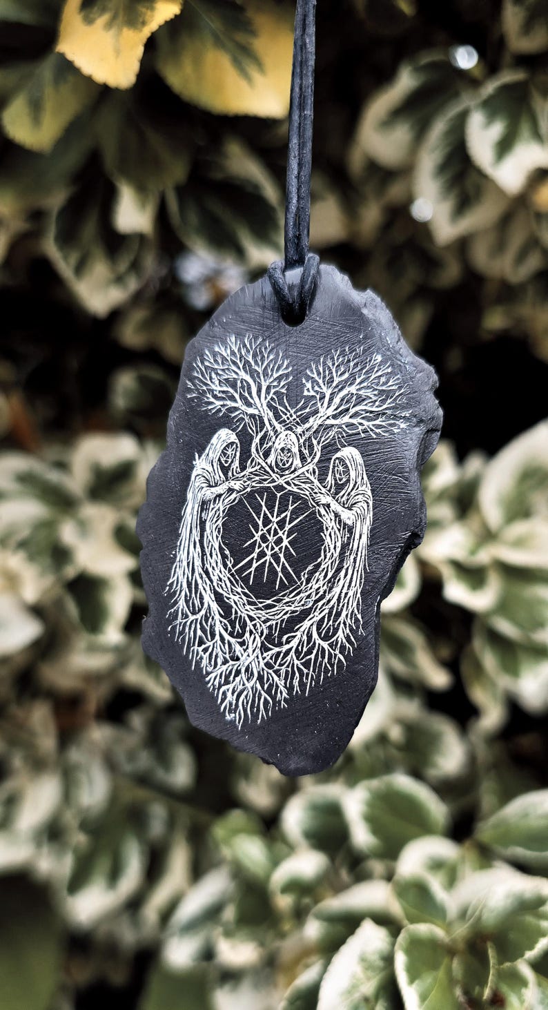 May include: A black slate pendant suspended on a dark cord, featuring a white engraved illustration of three hooded figures standing in a circle, their robes merging into intricate tree roots and branches surrounding a central geometric star symbol.