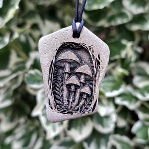 May include: A grey, irregularly shaped stone pendant featuring a detailed, dark-etched illustration of a cluster of mushrooms surrounded by foliage. The pendant is suspended from a thin, black cord through a hole at the top.