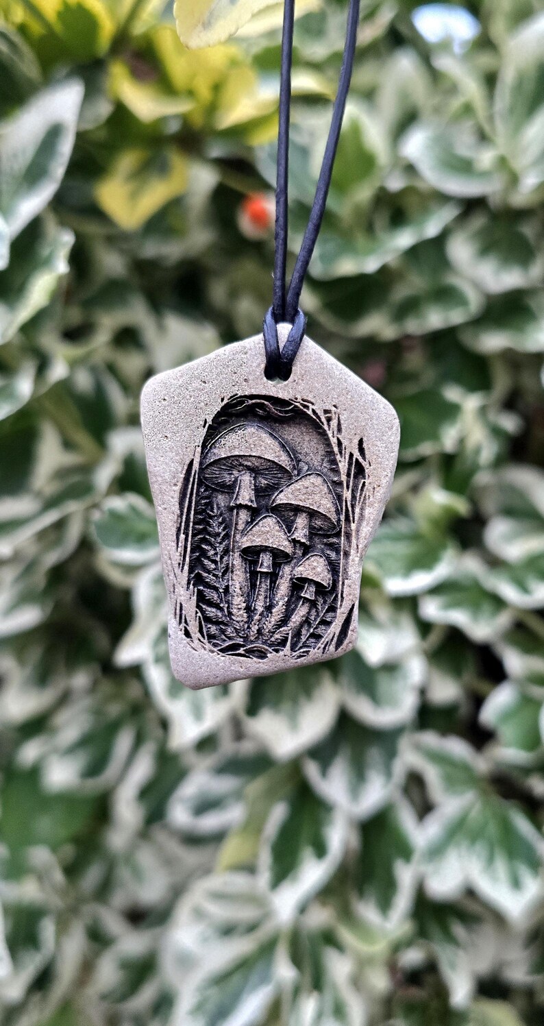 May include: A grey, irregularly shaped stone pendant featuring a detailed, dark-etched illustration of a cluster of mushrooms surrounded by foliage. The pendant is suspended from a thin, black cord through a hole at the top.