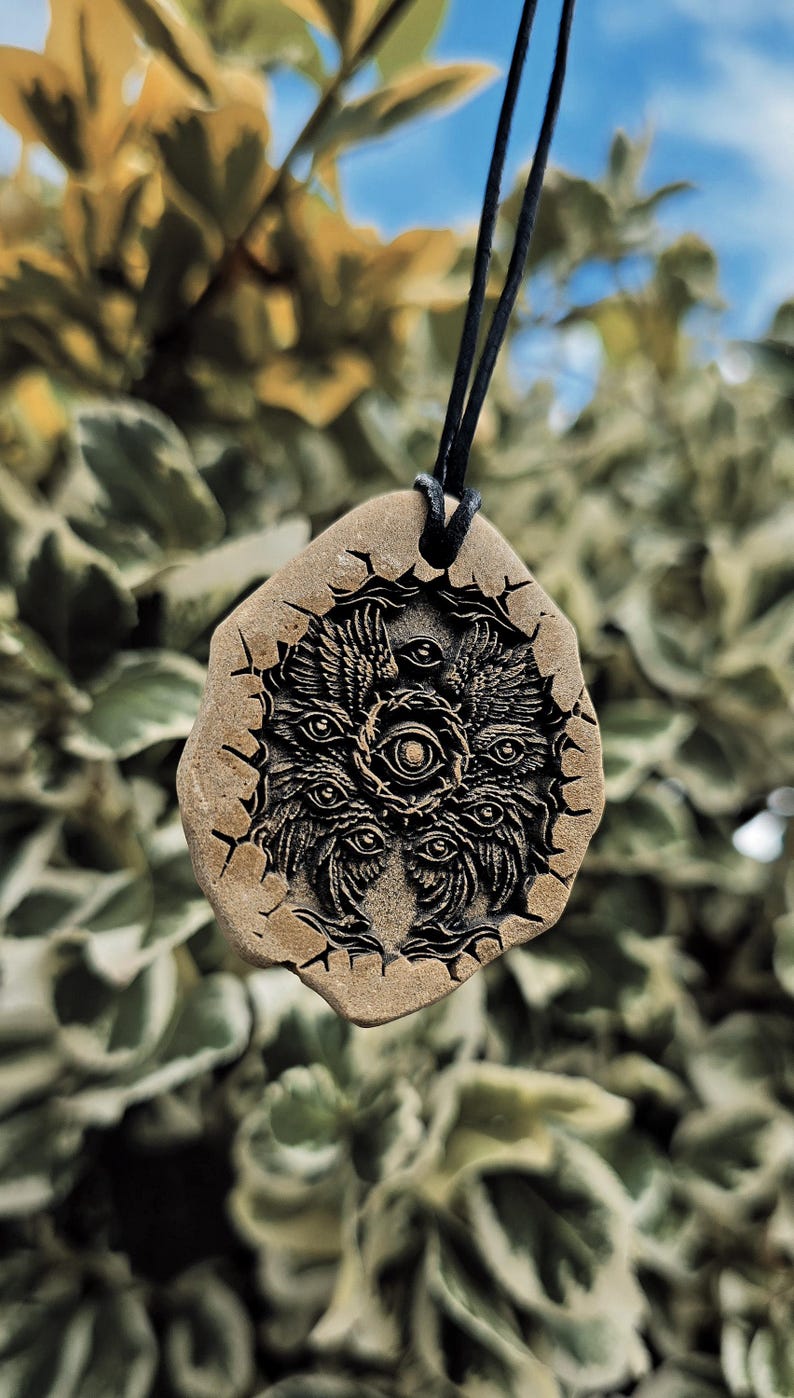 May include: A tan, irregularly shaped stone pendant suspended on a black cord. The surface features a deeply engraved, dark, intricate design of a multi-eyed, winged entity surrounded by a jagged, cracked circular border with radiating lines.