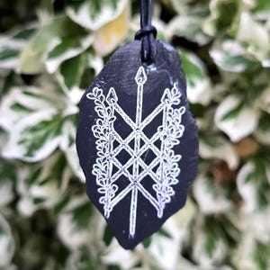 May include: A black, irregularly shaped stone pendant suspended from a black cord. The surface features a white, hand-drawn design of a central vertical arrow crossed by two diagonal arrows, flanked by intricate, leafy vine patterns on both sides.