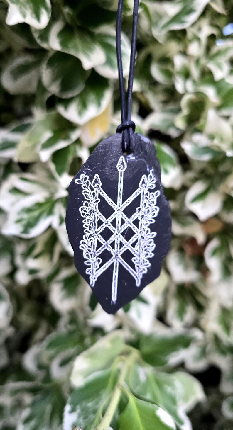 May include: A black, irregularly shaped stone pendant suspended from a black cord. The surface features a white, hand-drawn design of a central vertical arrow crossed by two diagonal arrows, flanked by intricate, leafy vine patterns on both sides.