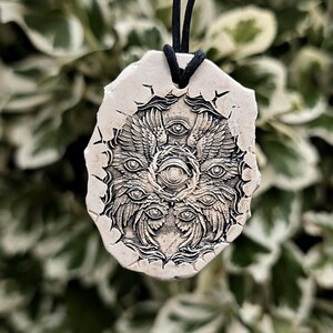 May include: A beige, irregularly shaped pendant featuring a detailed, dark-toned relief carving of a multi-winged, multi-eyed creature. The pendant is suspended from a thin black cord, showcasing a textured surface with intricate, symmetrical patterns.