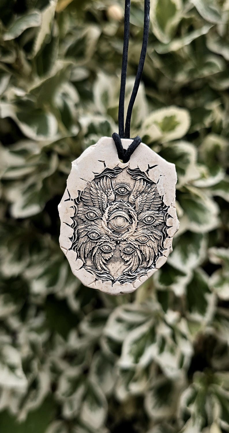 May include: A beige, irregularly shaped pendant featuring a detailed, dark-toned relief carving of a multi-winged, multi-eyed creature. The pendant is suspended from a thin black cord, showcasing a textured surface with intricate, symmetrical patterns.