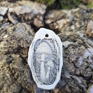 May include: An oval-shaped white pendant featuring a detailed, dark-toned relief carving of two mushrooms surrounded by fern-like foliage. The pendant has a small circular hole at the top for suspension and a smooth, irregular outer edge.