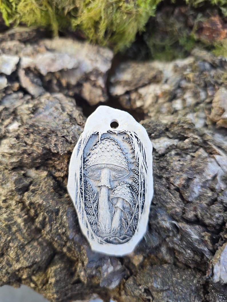 May include: An oval-shaped white pendant featuring a detailed, dark-toned relief carving of two mushrooms surrounded by fern-like foliage. The pendant has a small circular hole at the top for suspension and a smooth, irregular outer edge.