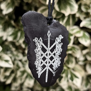 May include: A black, irregularly shaped stone pendant suspended from a black cord. The front surface features a white, hand-painted design of a central vertical spear-like symbol intertwined with a lattice pattern and leafy floral vines.