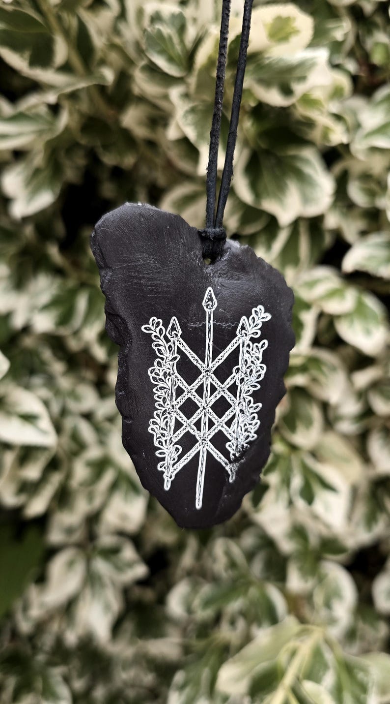 May include: A black, irregularly shaped stone pendant suspended from a black cord. The front surface features a white, hand-painted design of a central vertical spear-like symbol intertwined with a lattice pattern and leafy floral vines.