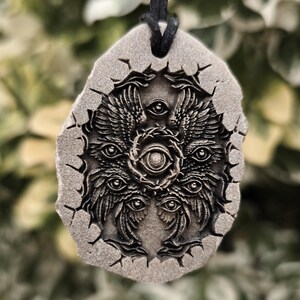 May include: A gray, stone-like pendant with a textured, cracked edge, suspended from a black cord. The surface features a detailed, relief-carved design of a multi-winged, multi-eyed entity with a central eye surrounded by a circular, thorny frame.