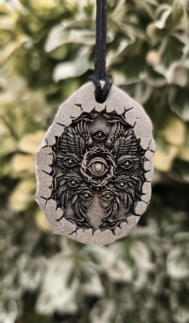 May include: A gray, stone-like pendant with a textured, cracked edge, suspended from a black cord. The surface features a detailed, relief-carved design of a multi-winged, multi-eyed entity with a central eye surrounded by a circular, thorny frame.