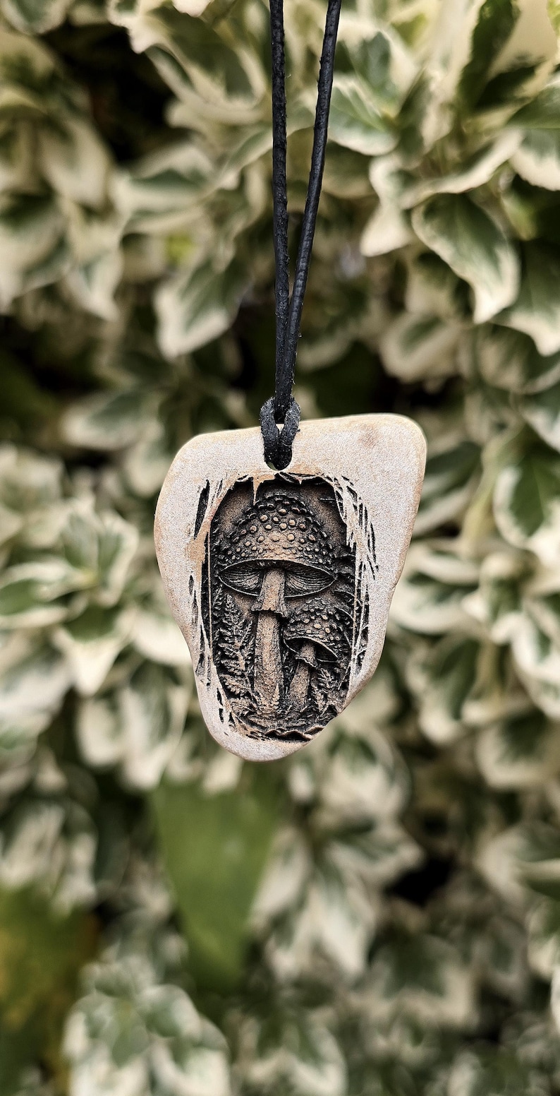 May include: A tan, irregularly shaped stone pendant suspended from a black cord. The centre of the stone features a detailed, dark, recessed carving of two mushrooms surrounded by ferns, creating a high-contrast, textured visual effect.