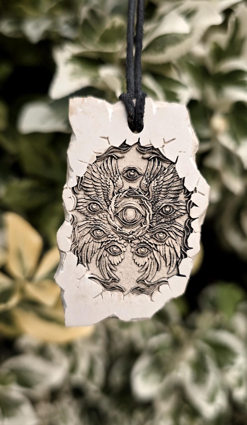 May include: A rectangular off-white stone pendant featuring a detailed dark engraving of a multi-eyed, winged celestial being surrounded by a crown of thorns. The pendant has a rough, uneven edge and is suspended from a simple black cord necklace.