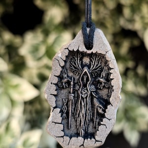 Puede incluir: Un colgante con un diseño detallado en gris oscuro de una figura encapuchada sosteniendo un bastón, enmarcado por una piedra agrietada de color gris claro. La figura está rodeada de pájaros estilizados y un elemento en forma de árbol. El colgante cuelga de un cordón negro.