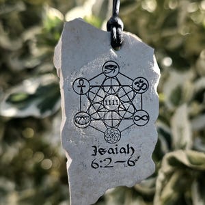 May include: A light gray, irregular stone pendant hanging from a black cord. The surface features a black printed design of a geometric Metatron's cube with various symbols, the number 1111, and the text Isaiah 6:2-6 printed in black ink.