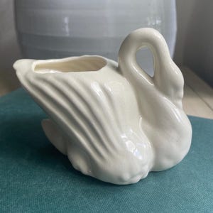 May include: A cream-colored ceramic swan-shaped planter. The swan has a curved neck and detailed wing feathers. The planter has a small opening for plants or flowers. The background includes a light-colored pot and a teal surface.