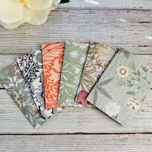 May include: A collection of six patterned notebooks fanned out on a wooden surface. The notebooks feature floral and botanical designs in various colors, including green, orange, and blue. The notebooks are rectangular and appear to be made of paper.