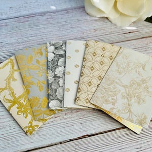 May include: A collection of six decorative paper envelopes with floral and geometric patterns in gold, gray, and cream. The envelopes are arranged in a fan shape, showcasing their various designs. The background is a light wood surface.