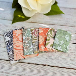 May include: A collection of folded paper cards with floral patterns in various colors, including blue, orange, and green. The cards are arranged in a fan shape on a light wood surface, with a white flower and green leaves in the background.