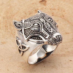 May include: A silver ring featuring a detailed wolf head design. The wolf's face is intricately carved with Celtic knotwork and geometric patterns.