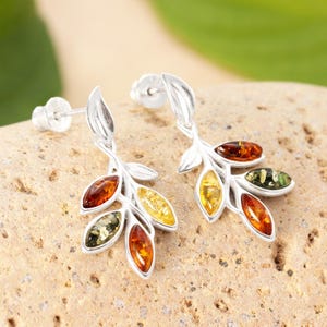 May include: Silver leaf earrings with amber-coloured stones. The earrings feature a leaf design with multiple amber stones in shades of orange, yellow, and green. The earrings are set on a neutral background.