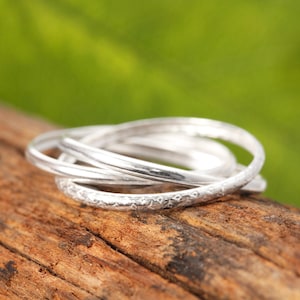 Puede incluir: Un conjunto de cuatro anillos de plata entrelazados. Uno tiene un diseño texturizado y martillado, los otros son lisos. Los anillos se muestran sobre un trozo de madera, con un fondo verde borroso.