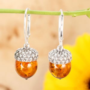 May include: A pair of silver acorn earrings with amber stones. The earrings are hanging from a green branch.