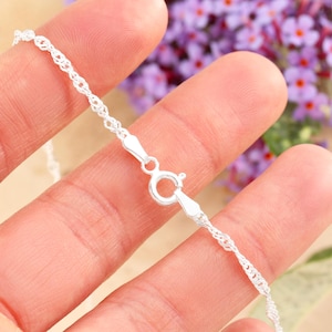 May include: Close-up of a delicate silver chain necklace with a lobster clasp. The chain has a twisted rope design, and the clasp is securely fastened. The background is blurred, with hints of purple flowers.