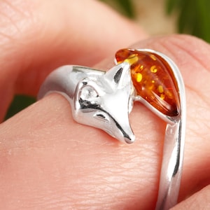 Könnte beinhalten: Silberner Fuch-Ring mit detailliertem Fuchskopf und tropfenförmigem Bernstein. Das Design des Rings weist eine polierte Oberfläche auf, die die Merkmale des Fuchses und die warmen Farbtöne des Bernsteins hervorhebt. Ein einzigartiges Schmuckstück.