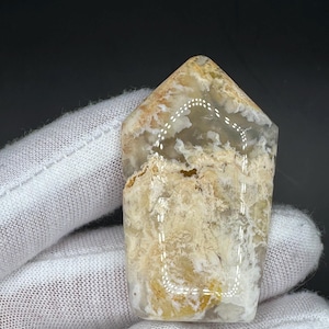 Puede incluir: Una piedra preciosa rectangular pulida con una parte superior redondeada. La piedra presenta una mezcla de tonos beige, crema y marrón claro con un aspecto nublado y texturizado. La gema es sostenida por una mano enguantada.