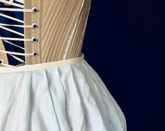 May include: A beige corset featuring diagonal stitching and silver metal grommets with white lacing, attached to a gathered light blue skirt. The garment is displayed on a form, showcasing the structured bodice and the soft, pleated fabric of the skirt.