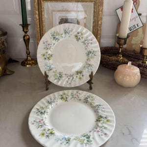 Könnte beinhalten: Zwei weiße Keramikteller mit einem Blumenmuster aus gelben, blauen und rosa Blüten. Die Teller haben einen gewellten Rand und werden auf einem dekorativen Ständer präsentiert. Sie sind Teil eines Vintage-Geschirrsets.