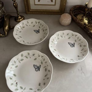 Puede incluir: Tres platos de cerámica blanca con bordes festoneados. Cada plato presenta un diseño de mariposa y hojas en gris y verde. Los platos están dispuestos sobre una superficie de mármol, lo que sugiere un tema de comedor o servicio.