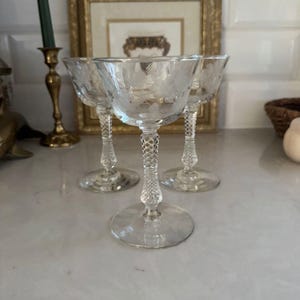 Könnte beinhalten: Drei klare, geätzte Champagner-Coupe-Gläser mit floralem Design. Die Gläser haben einen runden Fuß, einen strukturierten Stiel und eine flache Schale. Die Gläser werden auf einer hellen Oberfläche präsentiert.