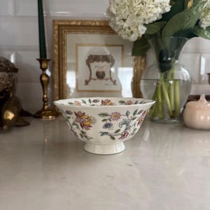 May include: A white porcelain bowl featuring a scalloped rim and a colorful floral pattern with pink, yellow, blue, and green botanical motifs. The bowl has a circular pedestal base and a smooth, glossy finish, showcasing intricate flower designs.