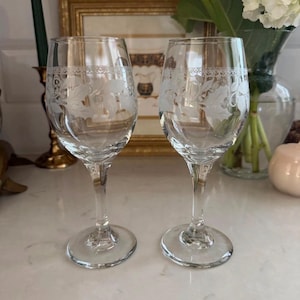 May include: Two clear glass wine glasses featuring a delicate etched pattern of leaves and vines encircling the upper portion of the bowls. Each glass has a smooth, transparent stem and a circular base, showcasing a refined and classic design.
