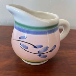 May include: A small, white ceramic creamer with a pink, blue, green and yellow striped design. The creamer has a floral design with blue leaves and a black stem.
