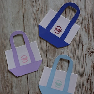 Pode incluir: Três pequenas bolsas de ombro com tecido branco e alças coloridas. As bolsas têm um logotipo que diz "Trading Junk" em um círculo. As bolsas são azuis, roxas e azul claro.