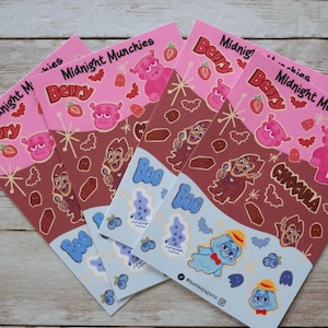 Puede incluir: Una colección de pegatinas rosas y marrones de Midnight Munchies Berry. Las pegatinas presentan personajes de dibujos animados, fresas y la palabra "Chocula". Pegatinas adicionales tienen fantasmas azules y un perro azul con sombrero.